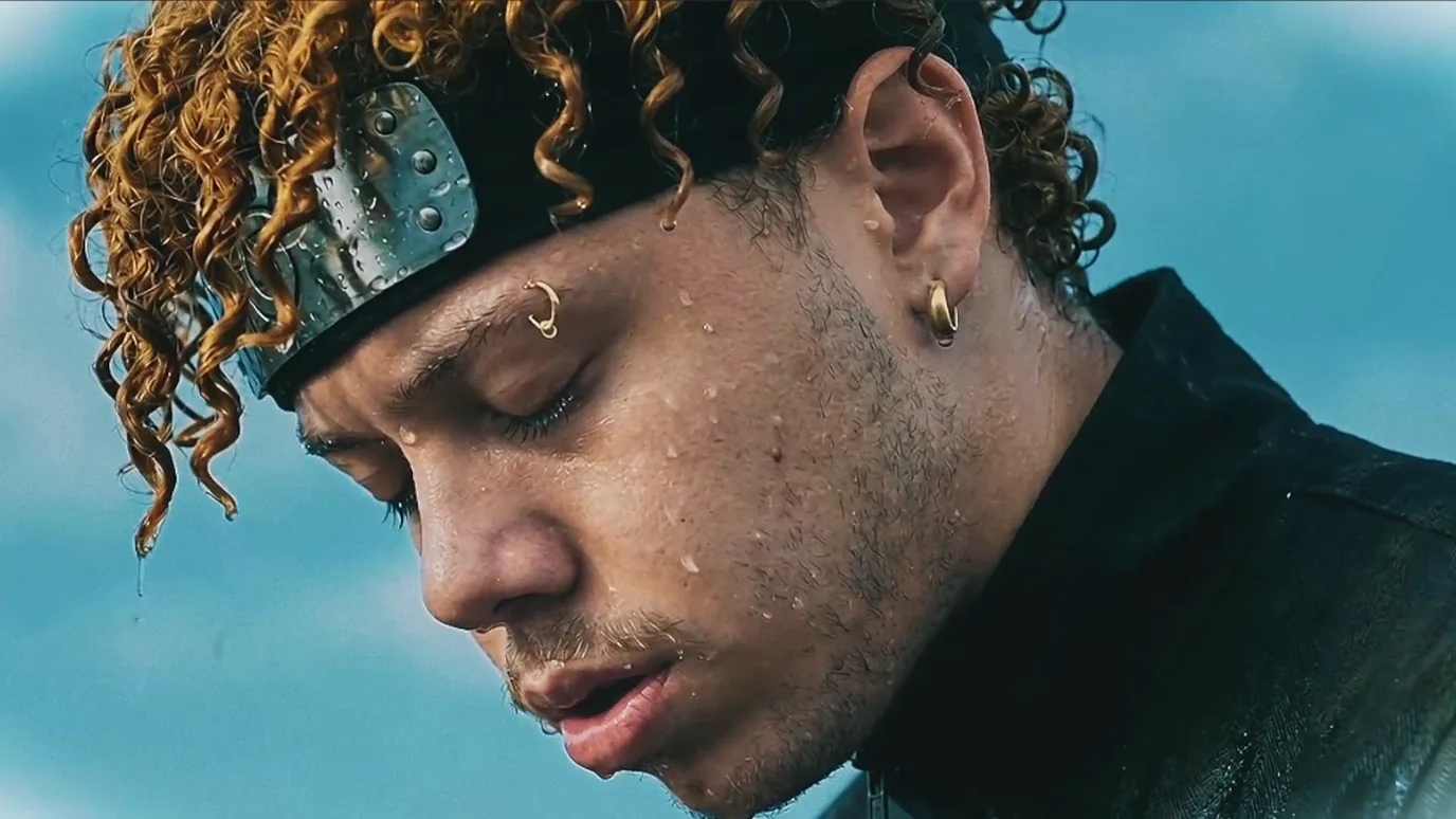 Extreme close on the man's face in cold daylight, head bowed, eyes closing, water running from his curls, pale sky behind him.