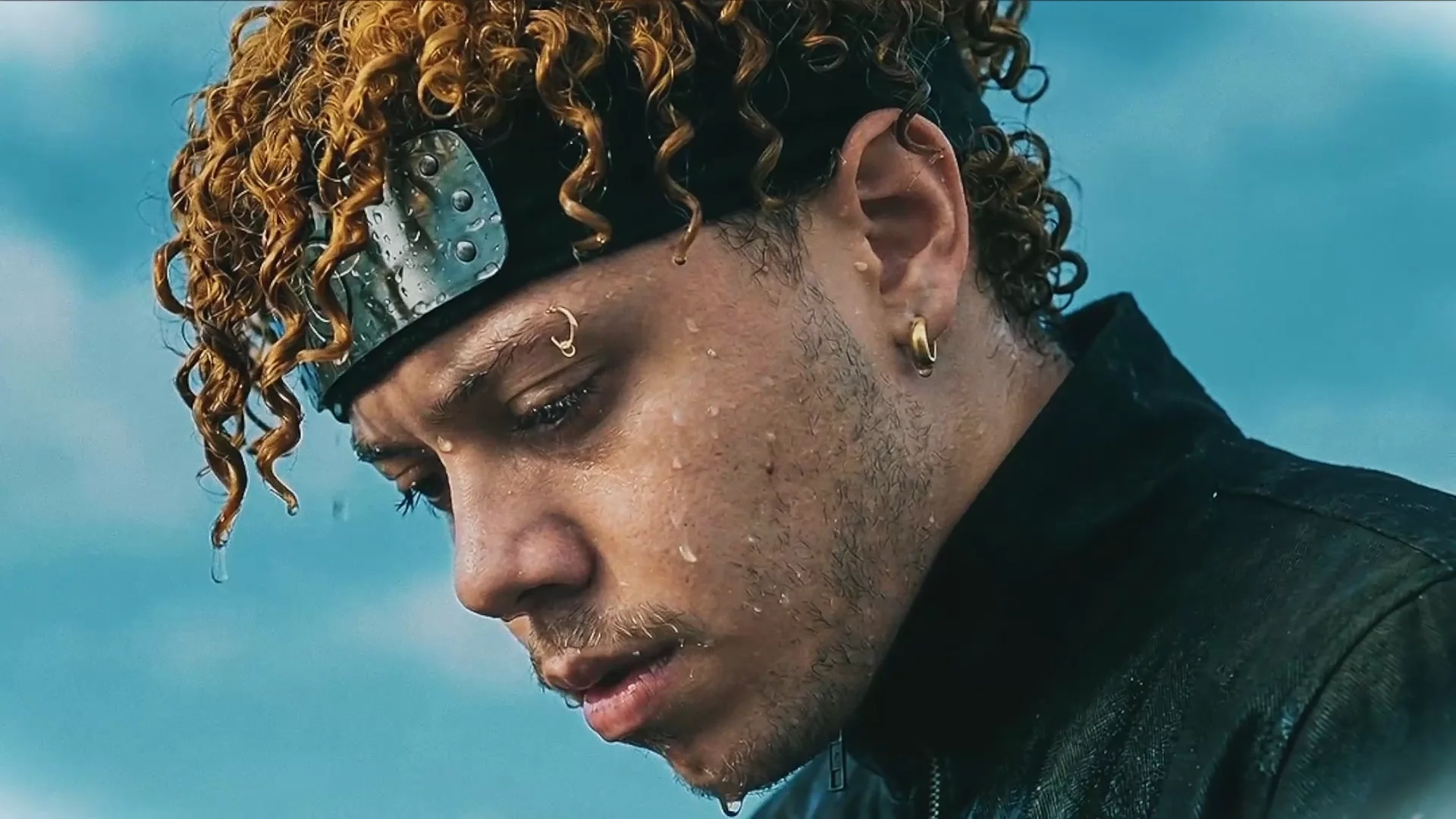 Close on a man's face in cold daylight, head bowed, eyes lowered, water running off his curls, a scratched steel plate on a black forehead band, pale sky behind him.